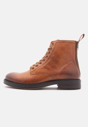 Bota de tobillo de cuero marrón con un acabado suave, cierre de cordones en la parte delantera, cremallera lateral y puntera redondeada. Suela plana de goma y detalles cosidos.