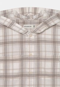 Beżowa koszula flanelowa w kratę z kapturem, z białymi i brązowymi paskami, dwiema przednimi kieszeniami i zapięciem na guziki. Metka wskazuje rozmiar 9/10.