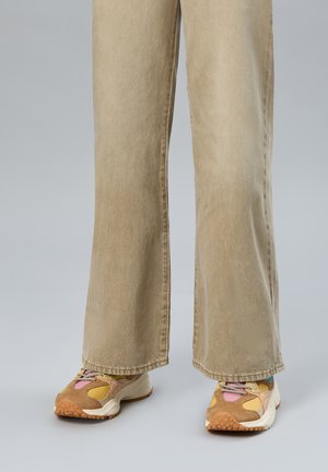 Jambes inférieures portant un pantalon large beige et des baskets multicolores avec des nuances de brun, jaune, rose et bleu sur un fond gris.