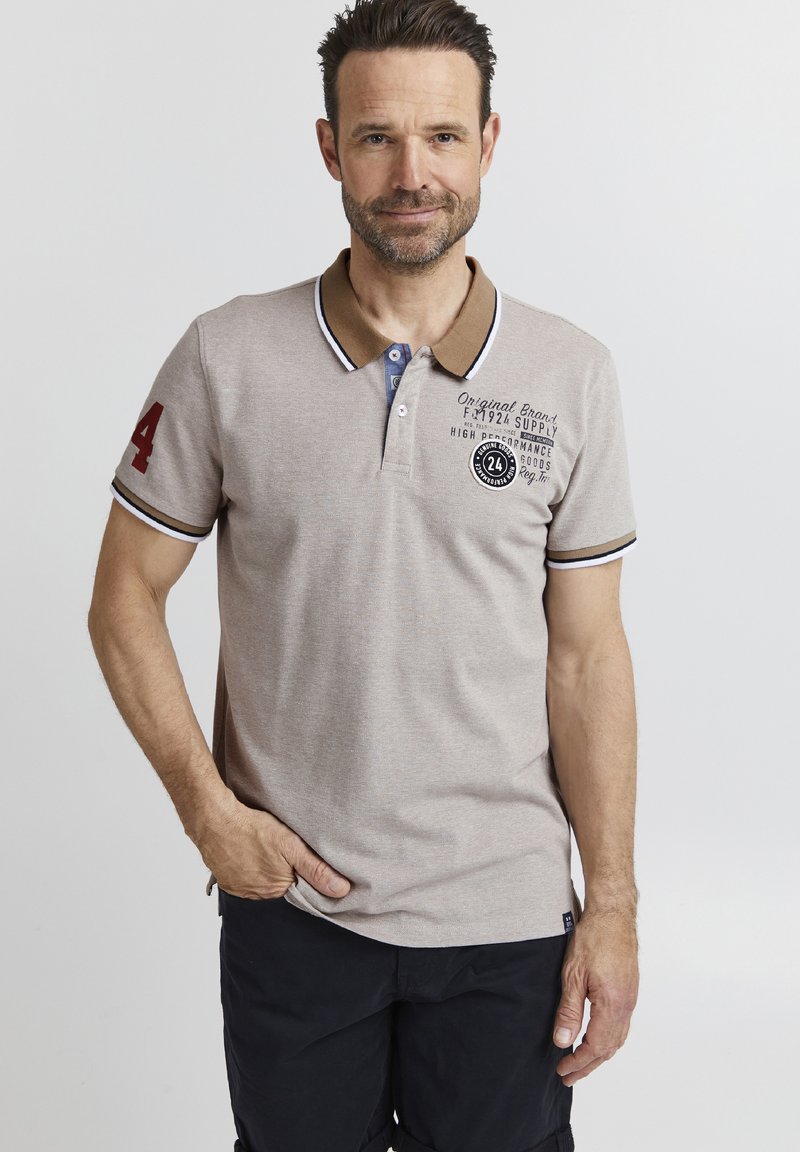 Camisa polo de color beige claro con cuello marrón. Presenta el número rojo "24" en la manga, botones de acento azules y detalles del logotipo impresos en negro.