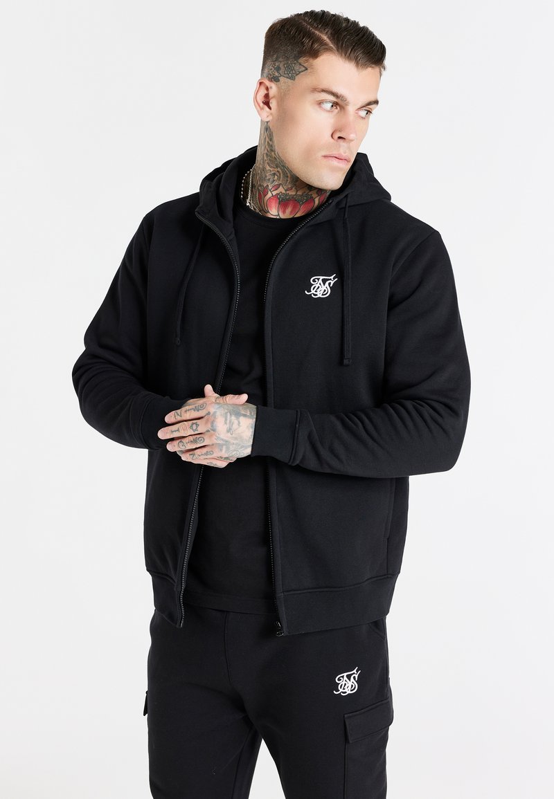 Black zip-up hoodie made of cotton blend, featuring a kangaroo pocket and logo embroidery on the chest. Smooth texture, adjustable hood.