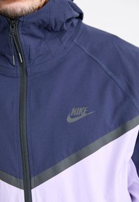 Veste Nike bleu marine et lavande avec un col montant, fermeture éclair et rayure d'accent grise ; présente un logo réfléchissant sur la poitrine.