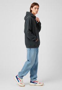 Donkergrijze hoodie met een voorzak, comfortabele pasvorm en verstelbare trekkoord. Gecombineerd met lichtblauwe spijkerbroeken en witte sneakers met rode en blauwe accenten.