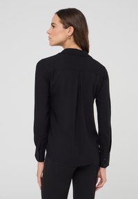 Blusa nera a maniche lunghe con colletto, polsini bottonati e design con pieghe sulla schiena. Tessuto liscio con silhouette aderente.