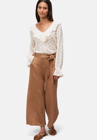 Blouse en dentelle avec un col et des manches à volants, associée à un pantalon large marron taille haute avec ceinture à nouer et poches. Ballerines marron.