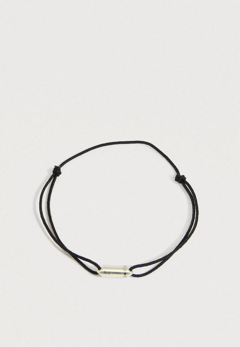 Crna narukvica s podesivom špagom i malim srebrnim cilindričnim privjeskom ugraviranim s "le gramme" i detaljima o težini.