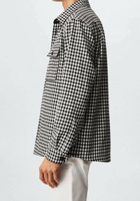 Homme portant une chemise à manches longues à motif pied-de-poule noir et blanc avec une poche poitrine, assortie à un pantalon de couleur claire, vu de profil.