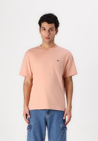 Tricou din bumbac roz deschis, cu mâneci scurte și guler rotund. Prezintă un mic logo verde Lacoste pe partea stângă a pieptului.
