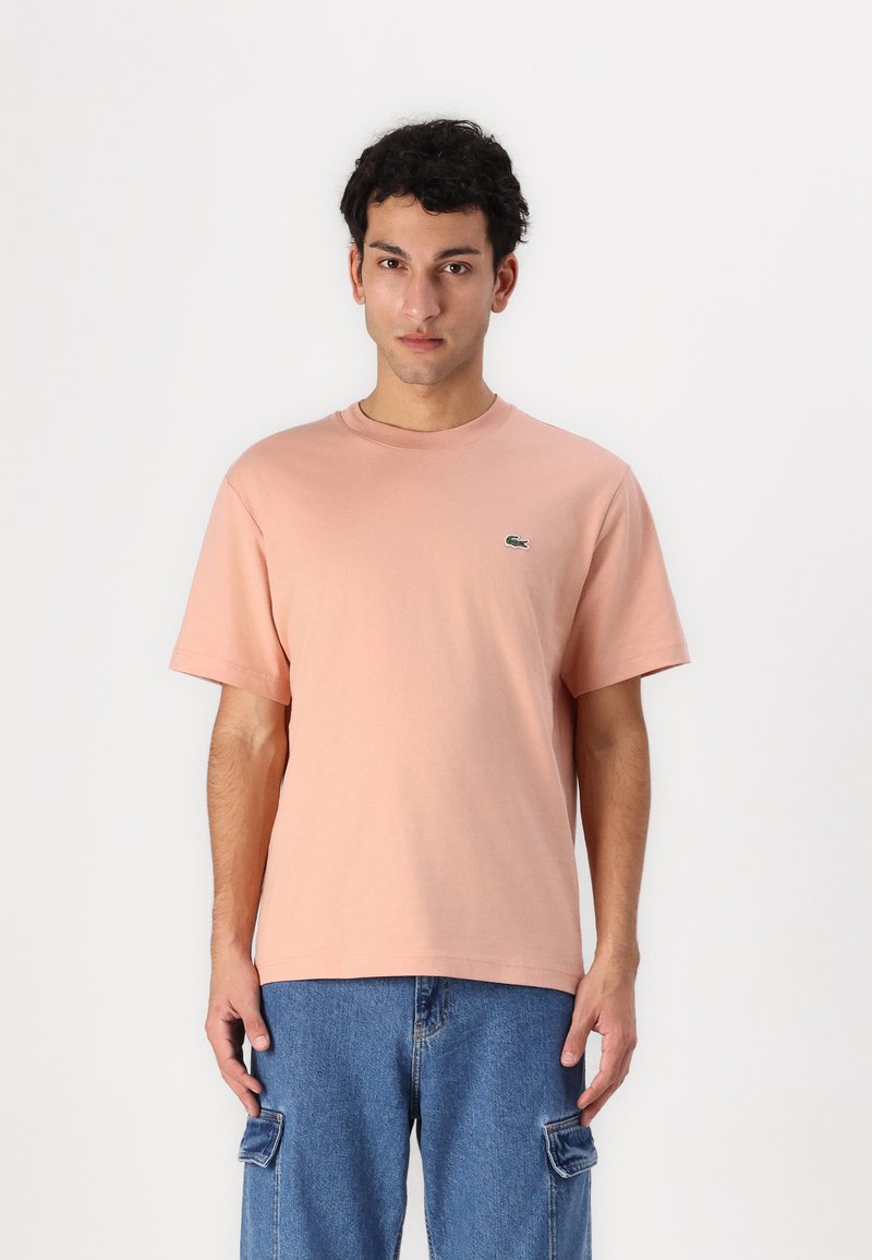 Tricou din bumbac roz deschis, cu mâneci scurte și guler rotund. Prezintă un mic logo verde Lacoste pe partea stângă a pieptului.