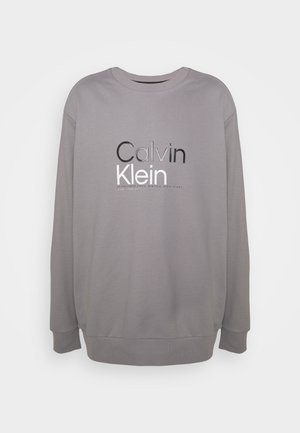 Szary sweter z okrągłym dekoltem, ze ściągaczami na rękawach i u dołu. Na przodzie znajduje się napis "Calvin Klein" w kontrastowych kolorach czerni i bieli.