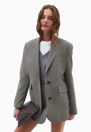 Karierter Oversized-Blazer aus grauem und schwarzem Stoff, mit zwei Knöpfen, Reverskragen und Fronttaschen, getragen über einem geschichteten grauen Outfit.