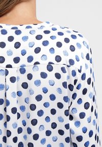 Tessuto bianco con pois blu e navy, caratterizzato da una vestibilità morbida, dettaglio con piega sul retro e una sottile texture.