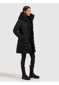 Kvinde iført lang sort quiltet frakke med høj krave, sort rullekrave, sorte læderbukser og sorte støvler, stående mod ensfarvet baggrund.