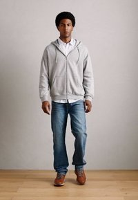 Un jeune homme portant un sweat à capuche gris, une chemise blanche, un jean bleu et des chaussures marron marchant sur un sol en bois devant un mur clair.