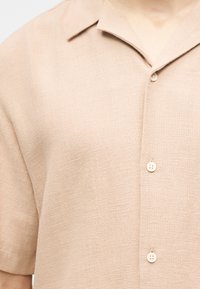 Μπεζ πλεκτό μπλουζάκι με κοντά μανίκια, γιακά τύπου spread και τρία εμφανή λευκά κουμπιά στο εμπρόσθιο μέρος.