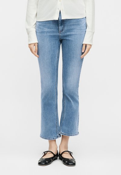 Άτομο που φοράει ανοιχτό μπλε cropped flare τζιν, λευκό πουκάμισο με μακριά μανίκια και μαύρα επίπεδα παπούτσια με ασημένια τρουκς.