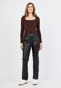 Zwarte leren broek met een aangesloten ontwerp en voorzakken, gecombineerd met een bruin langemouwen shirt. Model draagt witte sneakers.