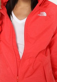 Veste zippée corail vif en tissu léger et texturé avec une capuche et un logo blanc sur la poitrine. Portée par-dessus une chemise blanche.