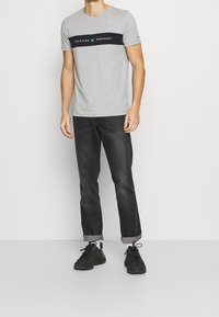 Camiseta de algodón gris con una franja horizontal negra que presenta texto, combinada con jeans oscuros y zapatillas negras.