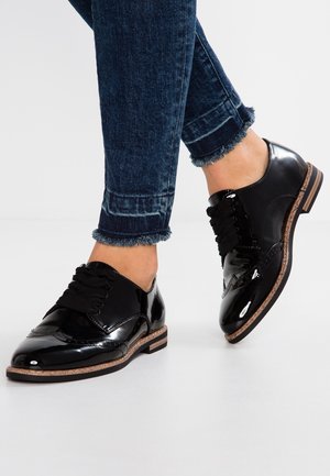 Chaussures en cuir verni noir avec un design à lacets, dotées de détails brogue et d'une semelle en liège naturel. Associées à des poignets en denim effilochés.