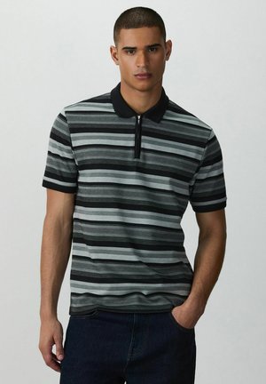 Man met kort haar, gekleed in een zwart-grijs gestreept polohemd met korte mouwen en een rits aan de voorkant, staande tegen een eenvoudige lichte achtergrond.