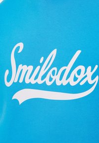 Camiseta de color azul claro con gran texto en cursiva blanca "Smilodox" y una textura suave de algodón. El diseño enfatiza el nombre de la marca de manera destacada.