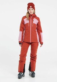 Rode en roze skijas met een ritssluiting aan de voorkant, contrasterende panelen, verstelbare capuchon, gecombineerd met bijpassende rode broek en zwarte bootsaccenten.