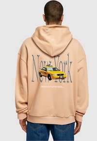 Beige kapucnis pulóvert viselő személy, a háta mögött nagy "New York" felirattal és sárga taxi grafikával, kék farmernadrággal, háttal a kamerának.