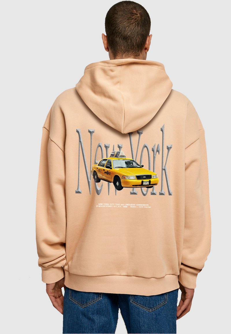 Beige kapucnis pulóvert viselő személy, a háta mögött nagy "New York" felirattal és sárga taxi grafikával, kék farmernadrággal, háttal a kamerának.