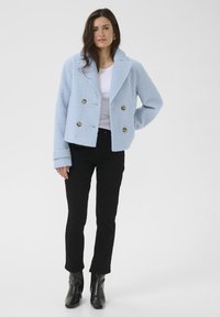 Veste double boutonnage bleu clair en tissu texturé avec des boutons dorés, portée sur un haut blanc, associée à un pantalon noir et des bottes élégantes.