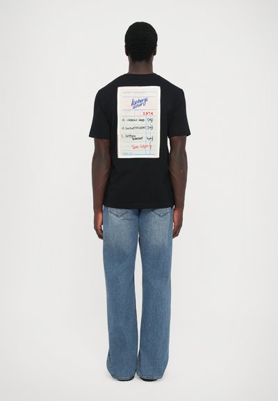 T-shirt noir avec un grand graphique d'un bloc-notes imprimé au dos, affichant des éléments de menu et un total. Porté avec un jean clair à jambe large.