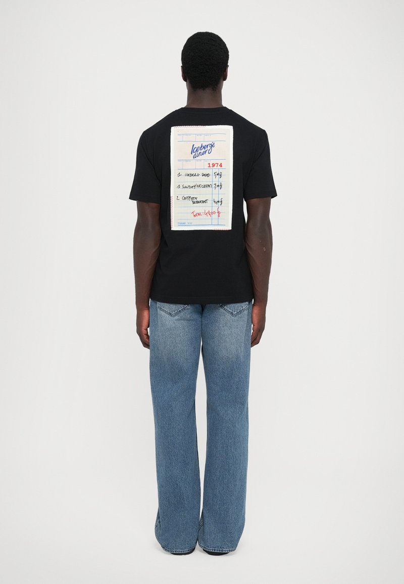 Schwarzes T-Shirt mit einer großen, bedruckten Notizblockgrafik auf der Rückseite, die Menüelemente und eine Gesamtsumme zeigt. Getragen mit hellblauen, weiten Jeans.