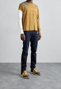 Camisa de manga larga mostaza con mangas blancas, jeans de mezclilla y zapatillas verdes con acentos caqui y una suela texturizada.