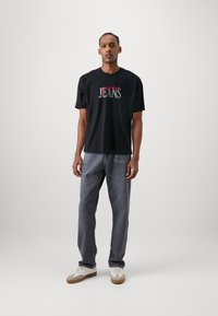 Camiseta negra de algodón con un texto enrojecido y gris estilizado que dice "Jeans". Combinada con jeans grises y zapatillas blancas. Diseño sencillo.
