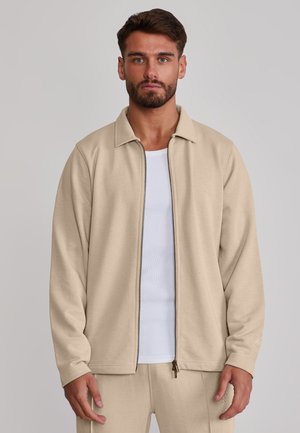 Mann trägt beige Reißverschlussjacke und passende Hose über weißem Rippshirt, steht vor einfarbigem grauem Hintergrund.