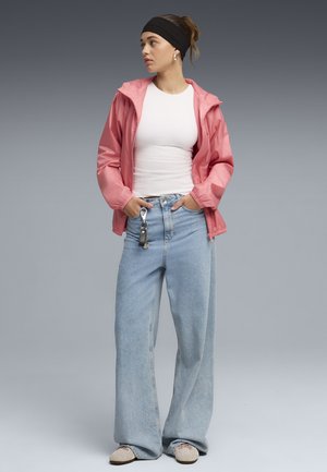 Ung kvinde iført en pink jakke, hvid top, vide lyseblå jeans og beige sneakers, stående med hænderne i lommerne og kigger mod venstre.