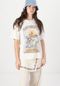 T-shirt crème avec un graphique de serpent coloré et un design de soleil ; associé à un pantalon beige clair avec broderie. Chapeau seau en bleu clair.
