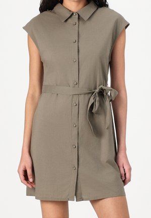 Ärmelloses khakifarbenes Hemdkleid mit Kragen und Stoffgürtel, der in einer Schleife an der Taille gebunden ist, getragen von einer Person mit entspannt an den Seiten hängenden Armen.