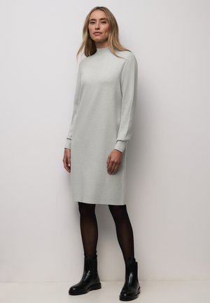 Frau steht an einer Wand, trägt ein langärmliges, hellgraues knielanges Kleid, schwarze Strumpfhosen und schwarze Stiefeletten.