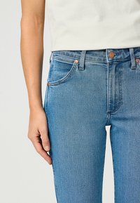 Jeans de tiro alto en azul, hechos de denim, con dos bolsillos delanteros, remaches de cobre y un acabado suave. Mano descansando sobre la pierna.