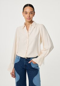 Crème knoopblouse met lange mouwen en parelversieringen bij de manchetten, gedragen met blauwe denim jeans met lichte vlekken.