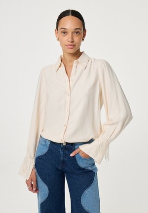 Crème knoopblouse met lange mouwen en parelversieringen bij de manchetten, gedragen met blauwe denim jeans met lichte vlekken.