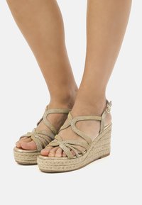 Sandales compensées avec une semelle en jute tissé, présentant des brides dorées métalliques dans un design entrecroisé, bout ouvert et une boucle réglable à la cheville.