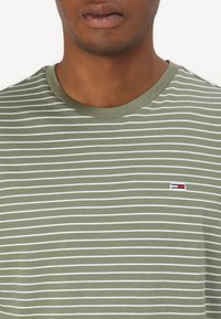 Camiseta de cuello redondo en verde oliva con rayas horizontales blancas. Pequeño logo con acentos en rojo, blanco y azul cerca del pecho. Material de algodón.