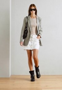 Blazer carreaux gris sur un haut beige, jupe en denim blanche avec des détails de boutons et un ourlet effiloché, bottines noires, avec un sac noir et des lunettes de soleil.
