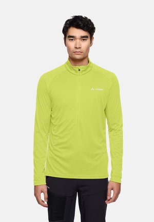 Mann trägt ein limegrünes, langärmliges Zip-up-Oberteil und schwarze Hosen, blickt nach vorne vor einem einfarbigen hellen Hintergrund.