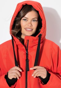 Veste imperméable rouge vif avec une capuche ajustée, des cordons de serrage réglables et une fermeture éclair noire. Présente des poignets élastiques et une texture lisse.