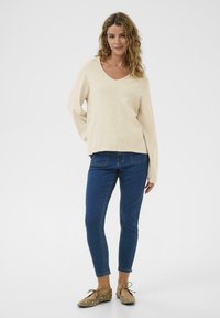 Beige Strickpullover mit V-Ausschnitt und lockerer Passform. Kombiniert mit enganliegenden blauen Jeans und hellbraunen Ballerinas mit weißen Akzenten. Glatte Textur und lässiges Design.