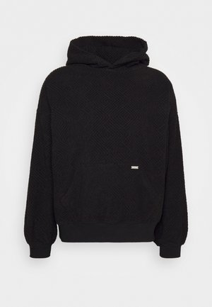 Personne portant un sweat à capuche noir texturé, un jean bleu clair et des baskets noires avec des semelles bleues, debout sur un sol bleu contre un mur blanc.
