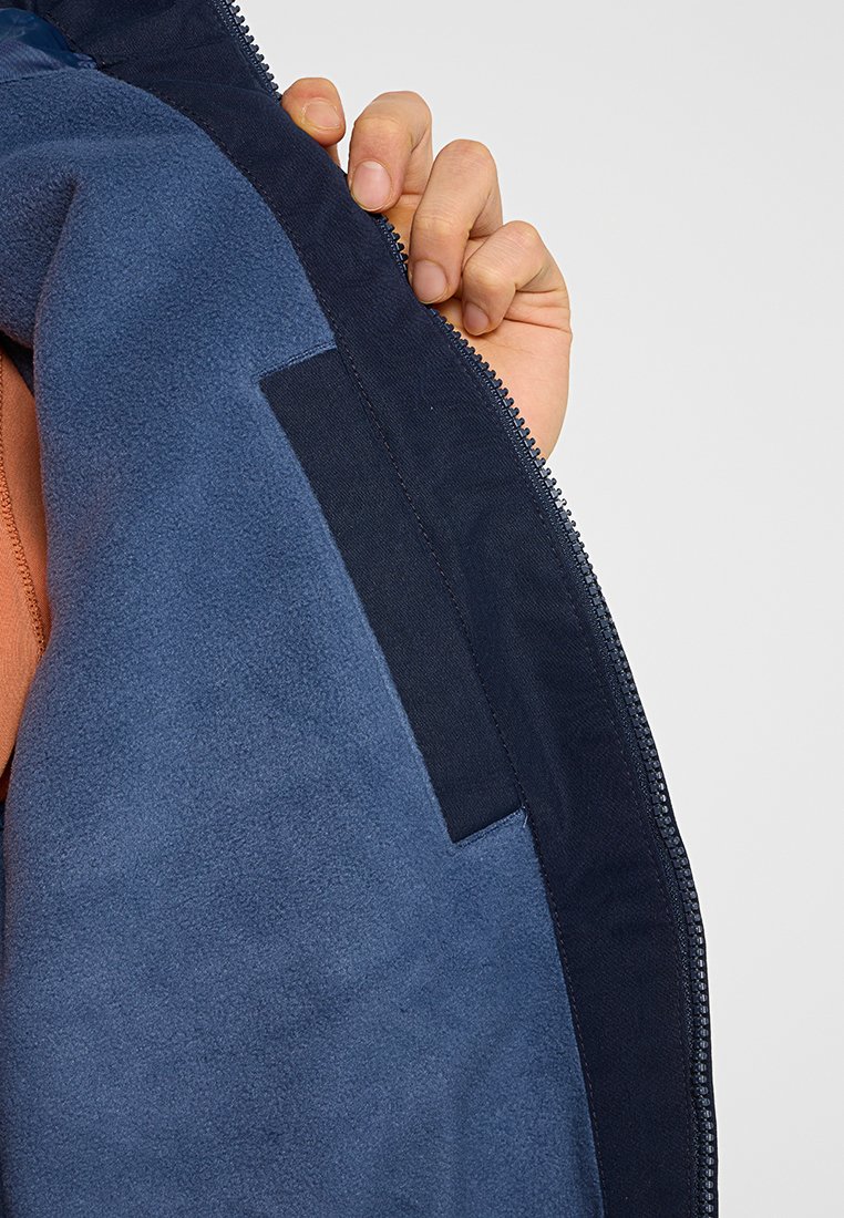 Ręka trzyma otwartą granatową polarową kurtkę, pokazującą wewnętrzny materiał i prostokątną wewnętrzną kieszeń.